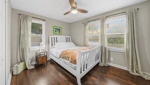 Quarto com uma cama branca, um ventilador de teto e duas janelas com cortinas estampadas.