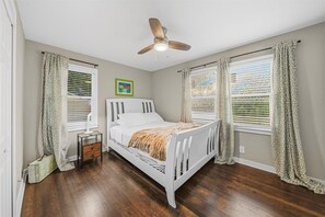Ein Schlafzimmer mit einem weißen Bett, einem Deckenventilator und zwei Fenstern mit gemusterten Vorhängen.