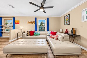 A living room with a sectional sofa, a ceiling fan, and a 'happy' sign on the wall. - Spacious 5BR Retreat w/ Game Room + Patio (Tampa)