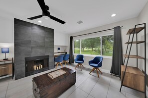 A modern living room with a fireplace, a ceiling fan, and a large window with a view of greenery. - Casa Solterra | 5 Bedroom Pool Retreat | Temple Terrace | Sleeps 12 (Temple Terrace)