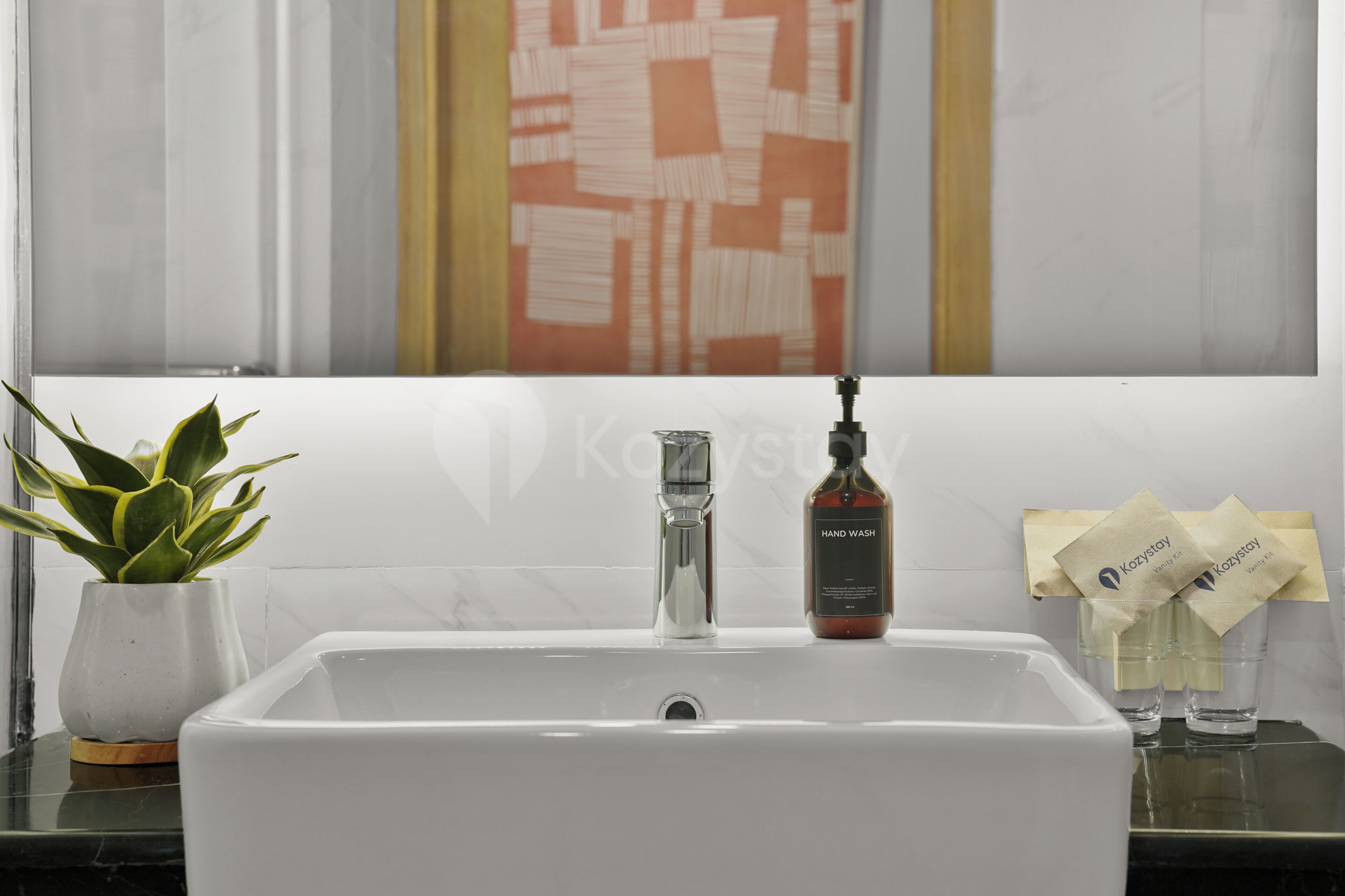 Un lavabo de salle de bain avec un robinet en verre transparent, une plante en pot, un flacon de savon liquide pour les mains et un distributeur de serviettes.