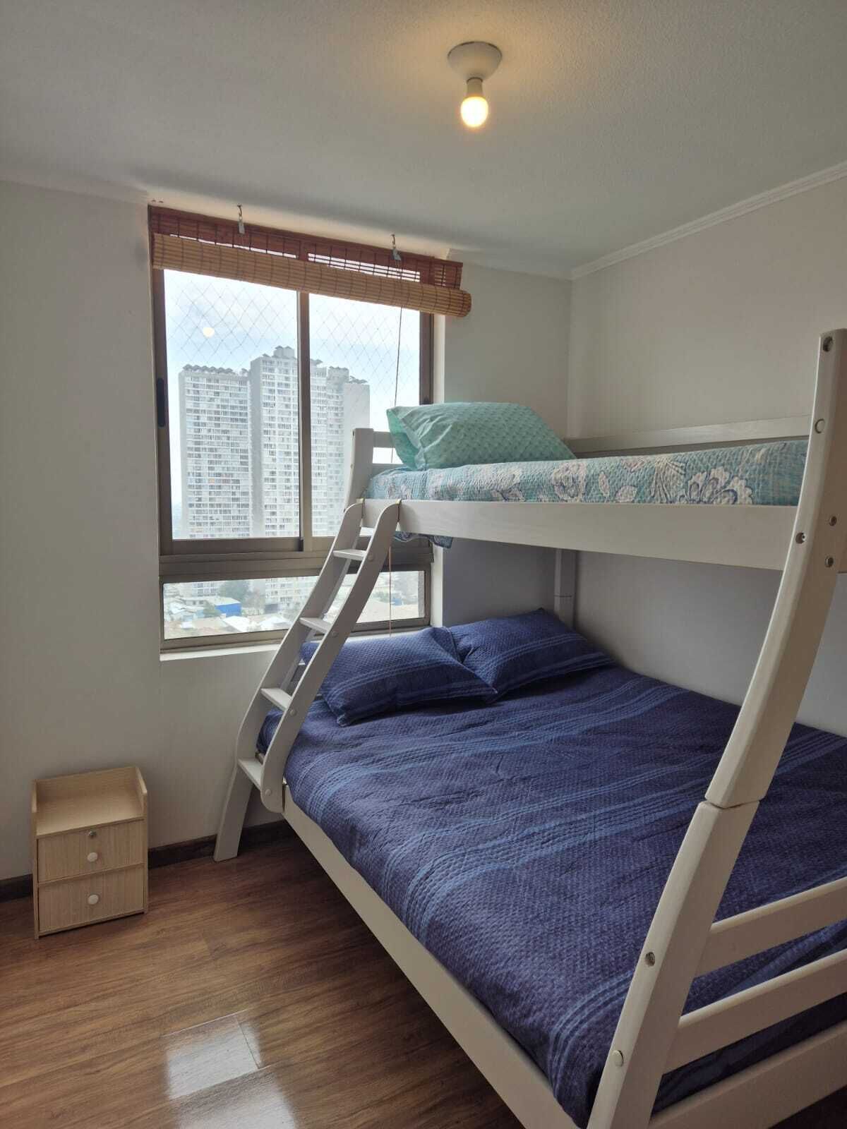 Dormitorio con literas, una ventana, un pequeño armario de madera y una lámpara en el techo.