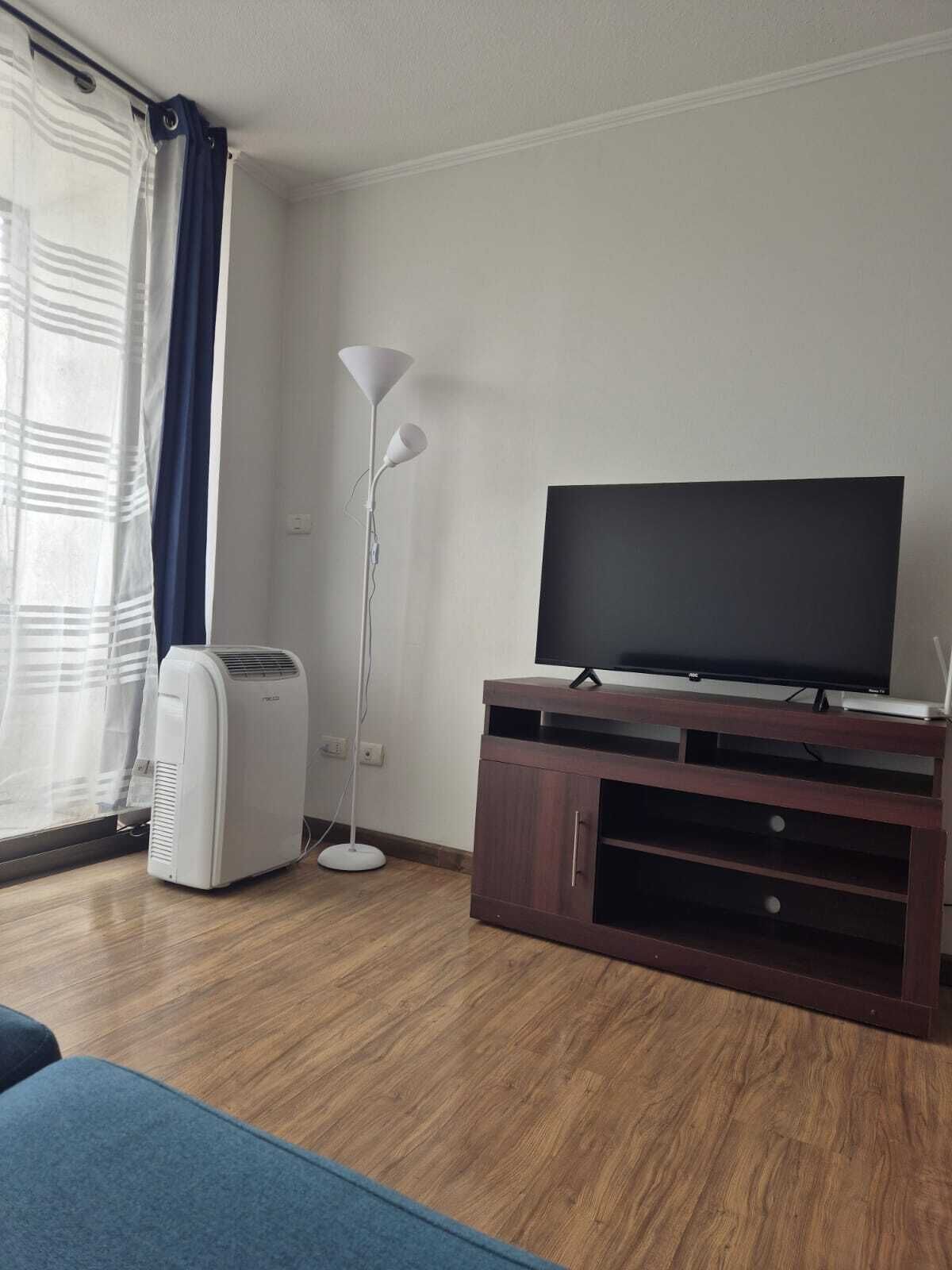 Una habitación con televisión de pantalla plana sobre un mueble de madera, un ventilador de pie y una ventana con cortinas.