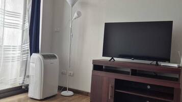 Una habitación con televisión de pantalla plana sobre un mueble de madera, un ventilador de pie y una ventana con cortinas.