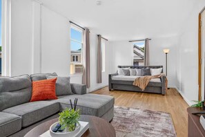 Ein modernes Wohnzimmer mit einem grauen Sofa, einem kleineren grauen Zweisitzer und einem Bett mit grau gestreuter Bettwäsche. Ein runder Couchtisch mit Pflanze und Fernbedienungen, ein gemustertes Teppich und eine Stehlampe sind ebenfalls vorhanden.