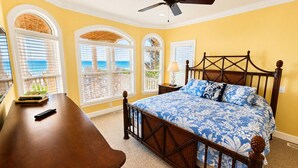 Ein Schlafzimmer mit einem Bett, einem Schreibtisch und einem Stuhl. Es gibt Fenster mit Jalousien, einen Deckenventilator und Meerblick.