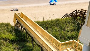 Eine Holzterrasse führt zu einem Strand mit Poolbereich und Liegestühlen.