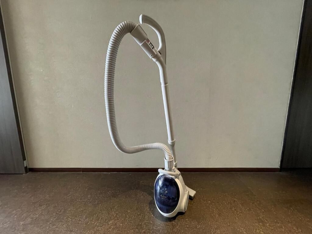 Un aspirateur muni d’un tuyau et d’un manche, posé sur le sol, contre un mur.