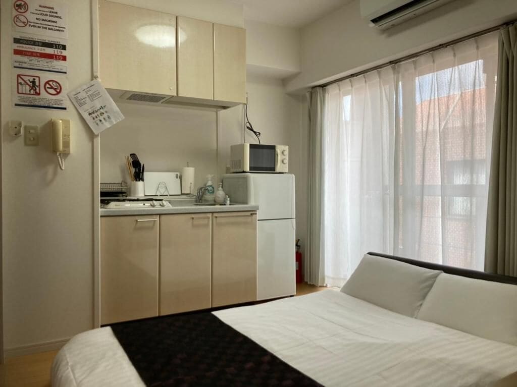 A hotel room with a bed, a kitchenette, a microwave, and a window with curtains.