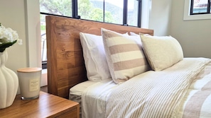 A neatly made bed with a wooden headboard, a bedside table with a candle, and a window with a view of greenery.