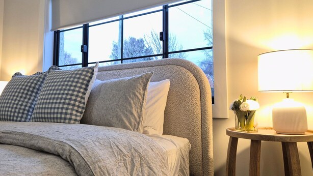 Ein Bett mit grauer Kopfteil, mehreren Kissen und einem Nachttisch mit Lampe und Blumenvase.