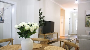 A living room with a television on a stand, a white vase with flowers on a table, and a sofa.