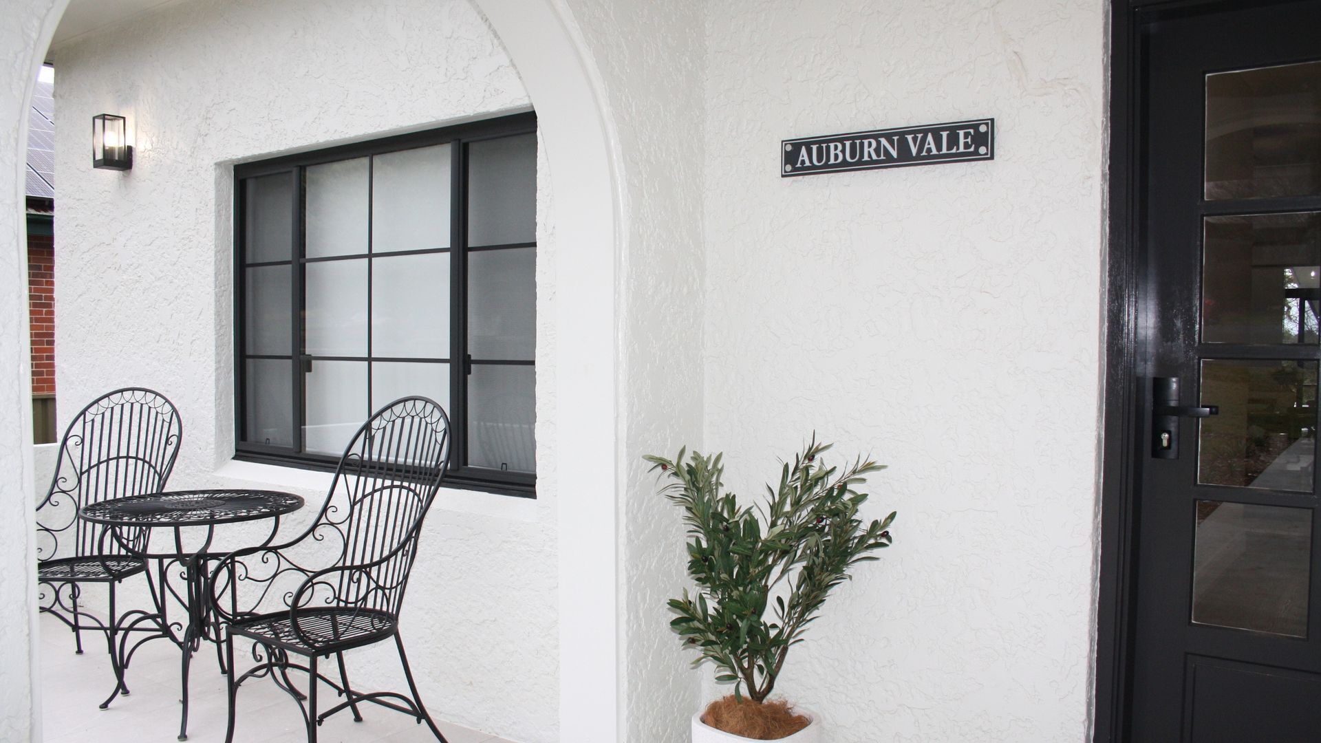A white building with a black door, a window with frosted glass, and a sign reading 'AUBURN VALE.' There are two metal chairs and a small potted plant.
