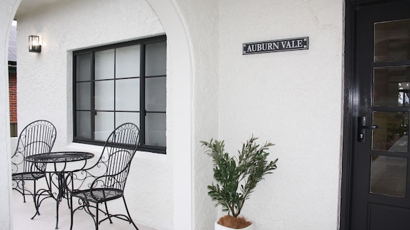 A white building with a black door, a window with frosted glass, and a sign reading 'AUBURN VALE.' There are two metal chairs and a small potted plant.