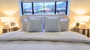 Ein Bett mit Kopfteil, zwei Nachttische mit Lampen und ein Fenster mit Blick auf Häuser und Autos.