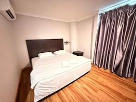 Un dormitorio con una cama grande, una mesita de noche, un aire acondicionado de pared y una ventana con cortinas.
