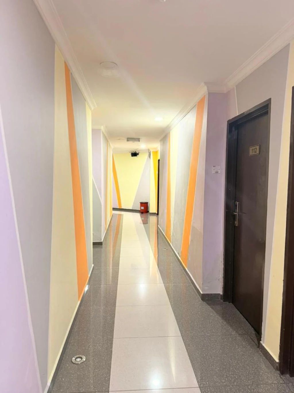 A hotel corridor with a tiled floor, a door with a number plate, and a trash bin.