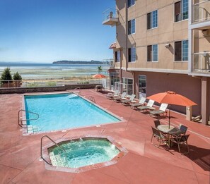 A rooftop pool with a hot tub, lounge chairs, and umbrellas, overlooking a scenic landscape.