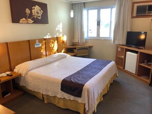 A hotel room with a large bed, a desk, a television, and a window with curtains. - Elegant Retreat with Modern Comforts & Peaceful Ambiance | Pub Station - 1.1 mi (Ponta Grossa)