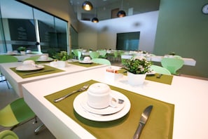 Une salle de conférence avec des tables préparées pour une réunion, équipée de chaises, d’assiettes, de couverts et de plantes en pot.