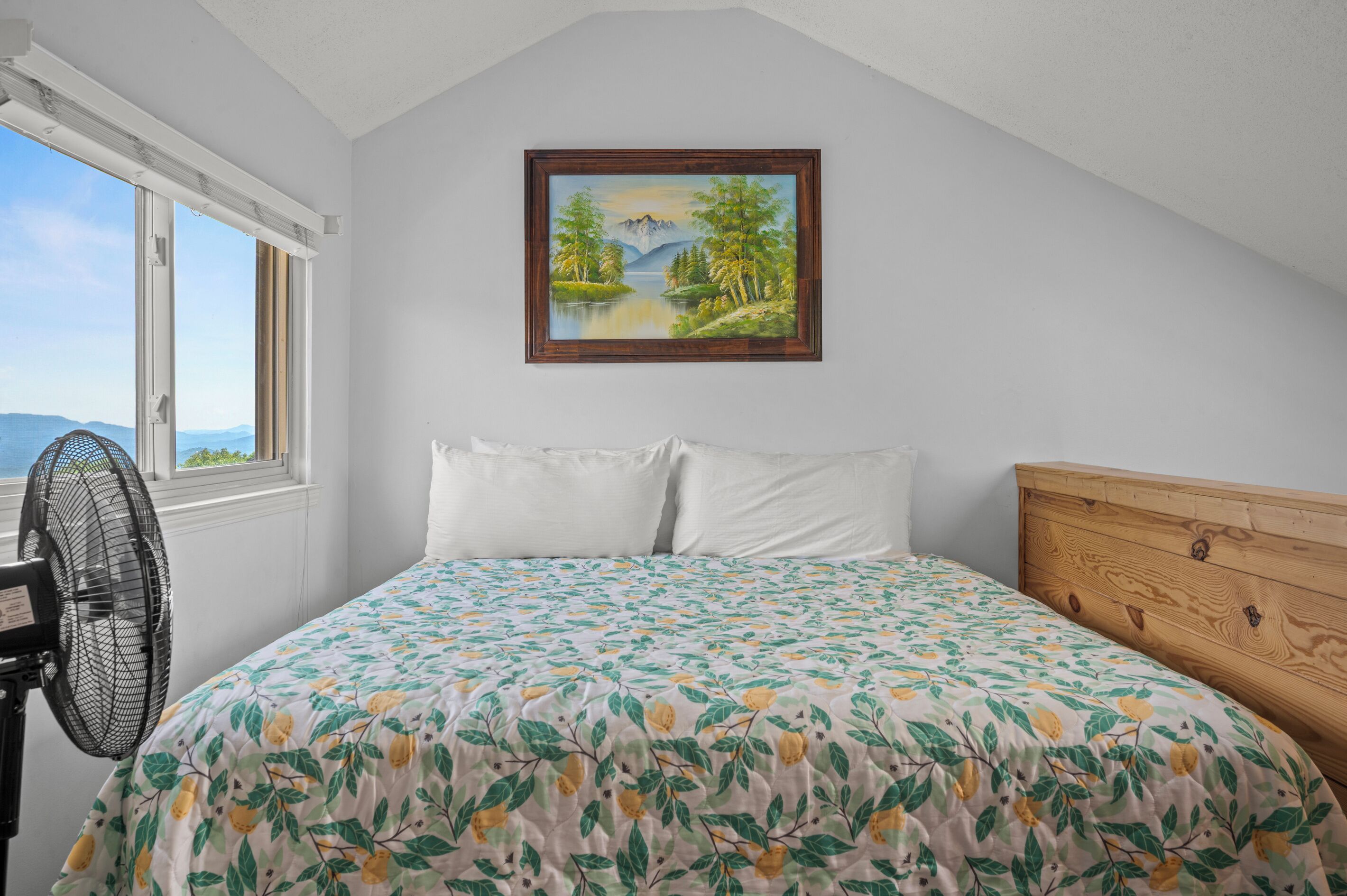 A bedroom with a bed, a window, a fan, and a painting on the wall.