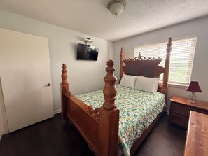 Ein Schlafzimmer mit Himmelbett, Wand-Fernseher, Fenster mit Jalousien und Nachttisch mit Lampe.