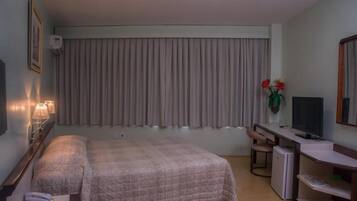 Quarto de hotel com cama, escrivaninha com computador, televisão e pequeno frigorífico.