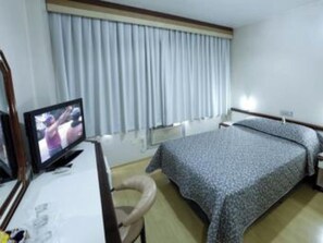 Ett hotellrum med en säng, ett skrivbord med en tv, en stol och ett fönster med gardiner.