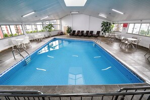 En inomhuspool med kristallklart blått vatten, omgiven av vita stolar och bord, och stora fönster som erbjuder utsikt över staden.
