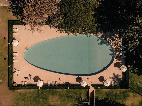 Luftaufnahme eines Schwimmbeckens mit Liegestühlen und Sonnenschirmen, umgeben von Bäumen.
