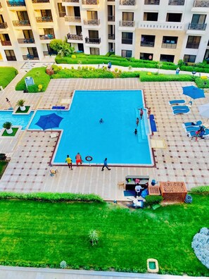 Ein Schwimmbecken, umgeben von einem Innenhof mit Schachbrettmuster, einigen Personen und einem Gebäude mit Balkonen im Hintergrund.