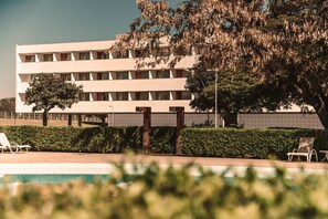 Un edificio moderno con piscina, sillas de descanso y un gran árbol en la fachada.
