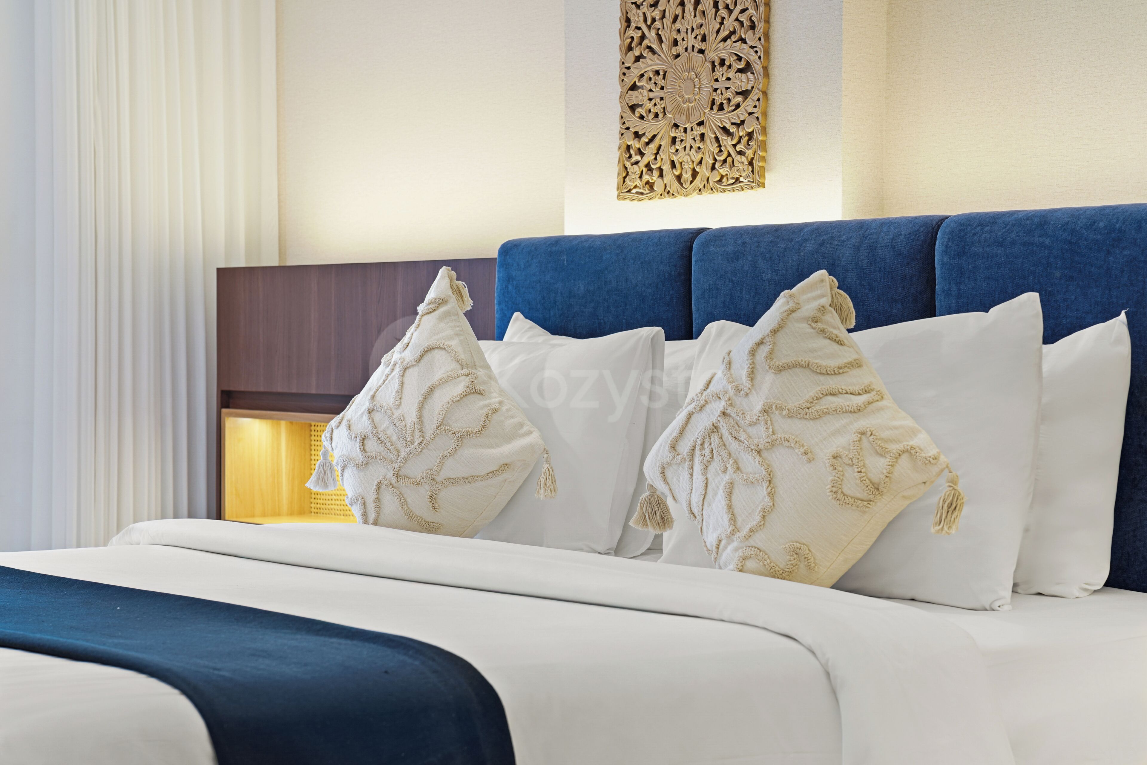 A hotel room with a neatly made bed, a blue headboard, and decorative pillows.