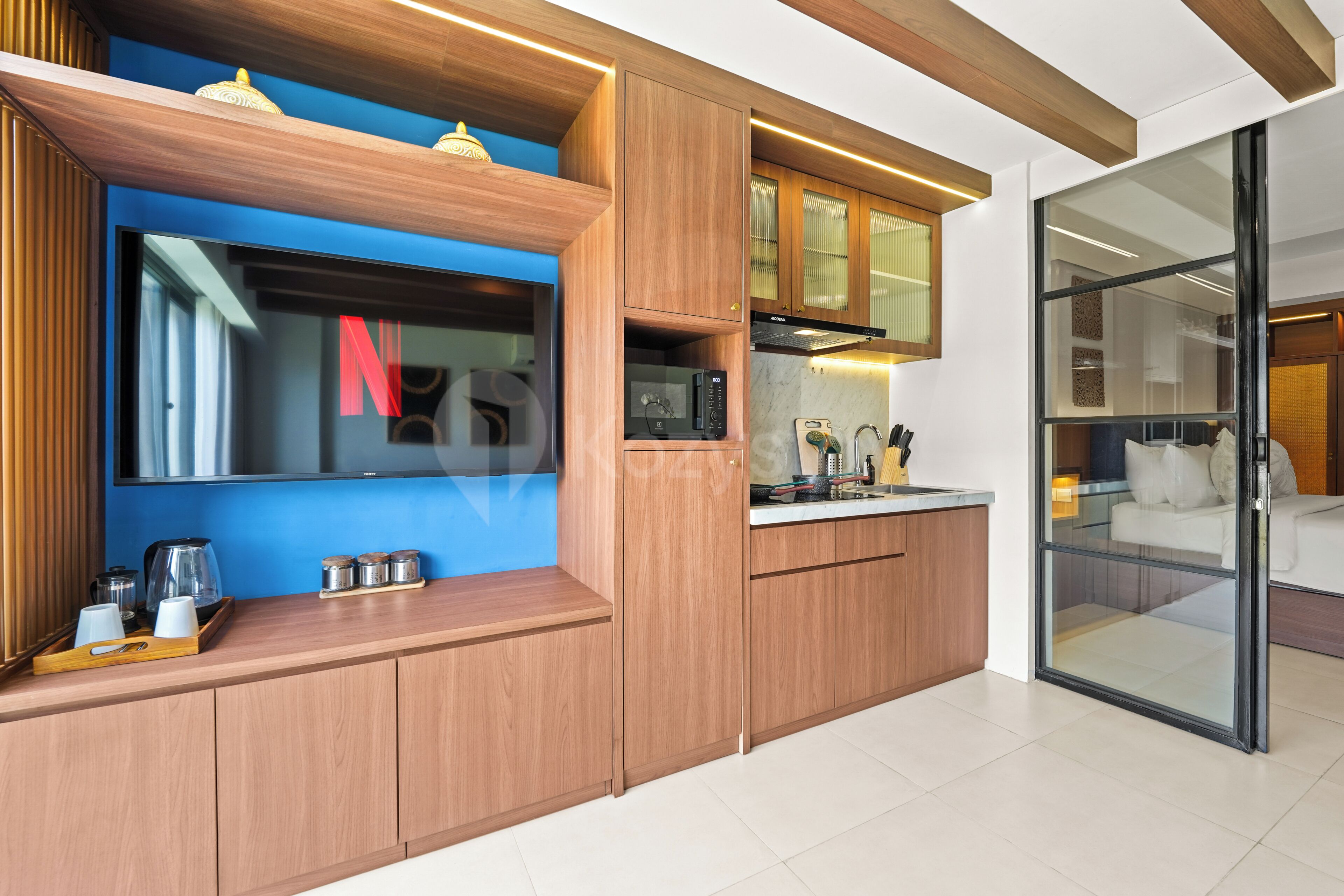 Een moderne keuken met houten kastjes, een ingebouwde magnetron en een glazen scheidingswand die naar een slaapkamer leidt.