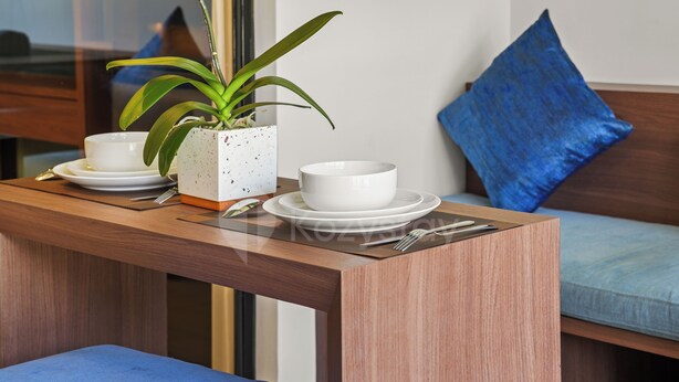 Un comedor con una mesa de madera preparada para dos personas, una maceta blanca con una planta y un cojín azul en un sofá.