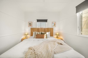 Ein Schlafzimmer mit einem Bett, zwei Nachttischlampen, einem Fenster mit Jalousien und zwei gerahmten Bildern an der Wand.