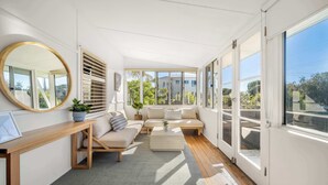 Um quarto claro e iluminado pelo sol, com uma janela grande, um espelho redondo, um sofá, uma mesinha e um vaso com planta.