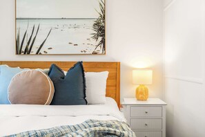 Ein ordentlich bezogenes Bett mit Kissen, einer Nachttischlampe und einem gerahmten Bild mit Strandmotiv an der Wand.