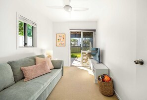 A living room with a grey sofa, a television on a stand, a ceiling fan, and a window with blinds. - Aloha Sands - Golden Beach Escape, Dog-Friendly (Caloundra Region)