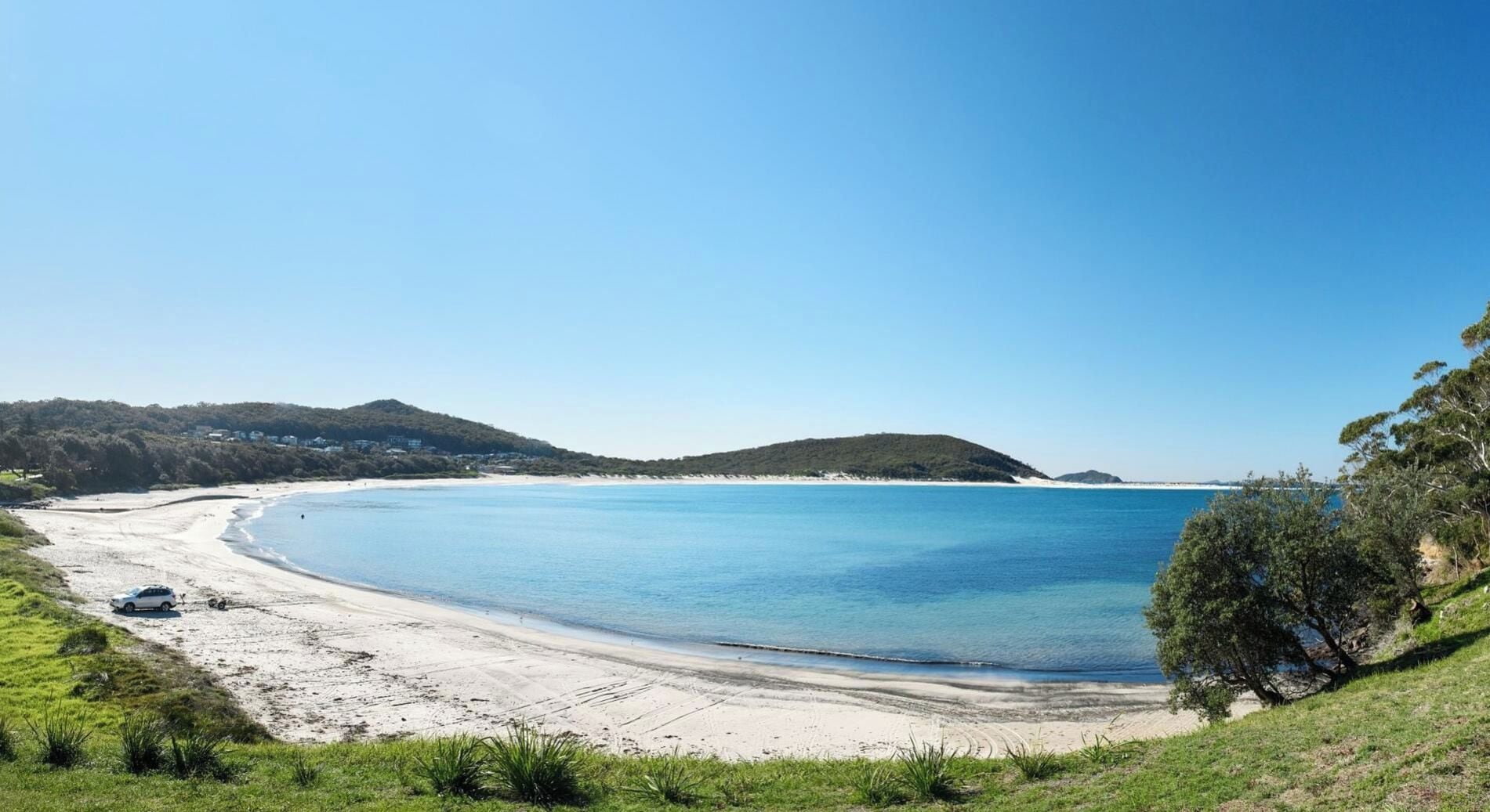 A coastal landscape with a sandy beach, clear blue water, and green hills in the background.
