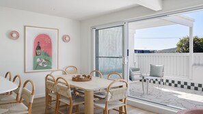 A dining area with a table and chairs, a framed picture on the wall, and a view of the outside balcony. - Ornos 3 Bedroom Apartment in Fingal Bay (Fingal Bay)