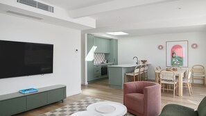 Ein modernes Wohnzimmer mit Flachbildfernseher, einem grünen Sofa, einem rosafarbenen Sessel und einem Essbereich mit Tisch und Stühlen.