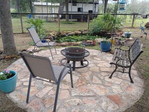 Eine Terrassenfläche im Freien mit Steinplatten, zwei Metallstühlen, einer runden Feuerstelle und Kübelpflanzen.