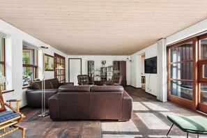 A spacious living room with a large brown sofa, a wooden floor, and a large window with curtains. - Holiday home with billiards, shelter & fire pit (Silkeborg)
