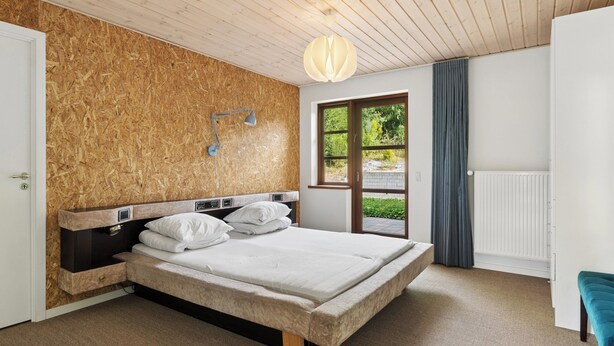 A bedroom with a wooden headboard, a bed with white bedding, a bedside lamp, a window with a view of greenery, and a radiator. - Holiday home with billiards, shelter & fire pit (Silkeborg)