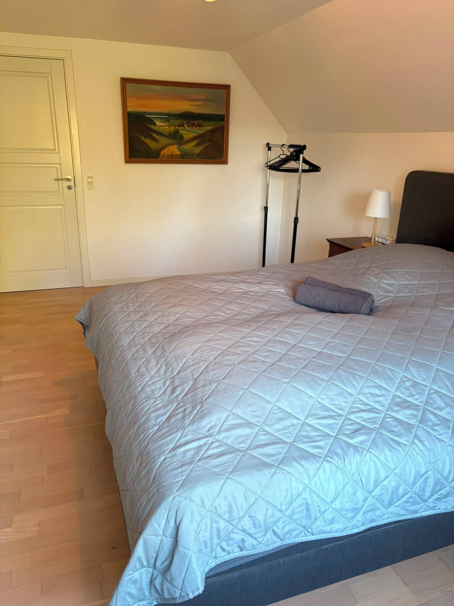 A bedroom with a large bed, a painting on the wall, a chair, and a lamp.