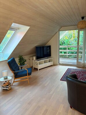 A loft-style room with a sloped ceiling, a television on a white entertainment unit, a blue chair, a black sofa, and a view of greenery through large windows. - Scenic home in Virklund (Virklund)