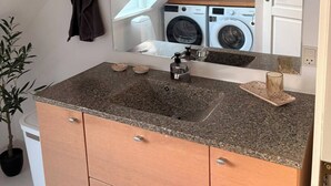 A bathroom with a granite countertop, a sink, a mirror, a towel rack, a potted plant, and a washing machine in the background. - Scenic home in Virklund (Virklund)