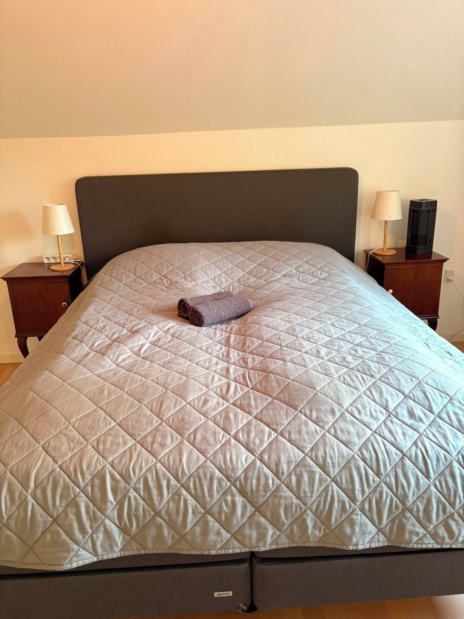 A bed with a quilted headboard, a rolled towel, and two bedside tables with lamps.