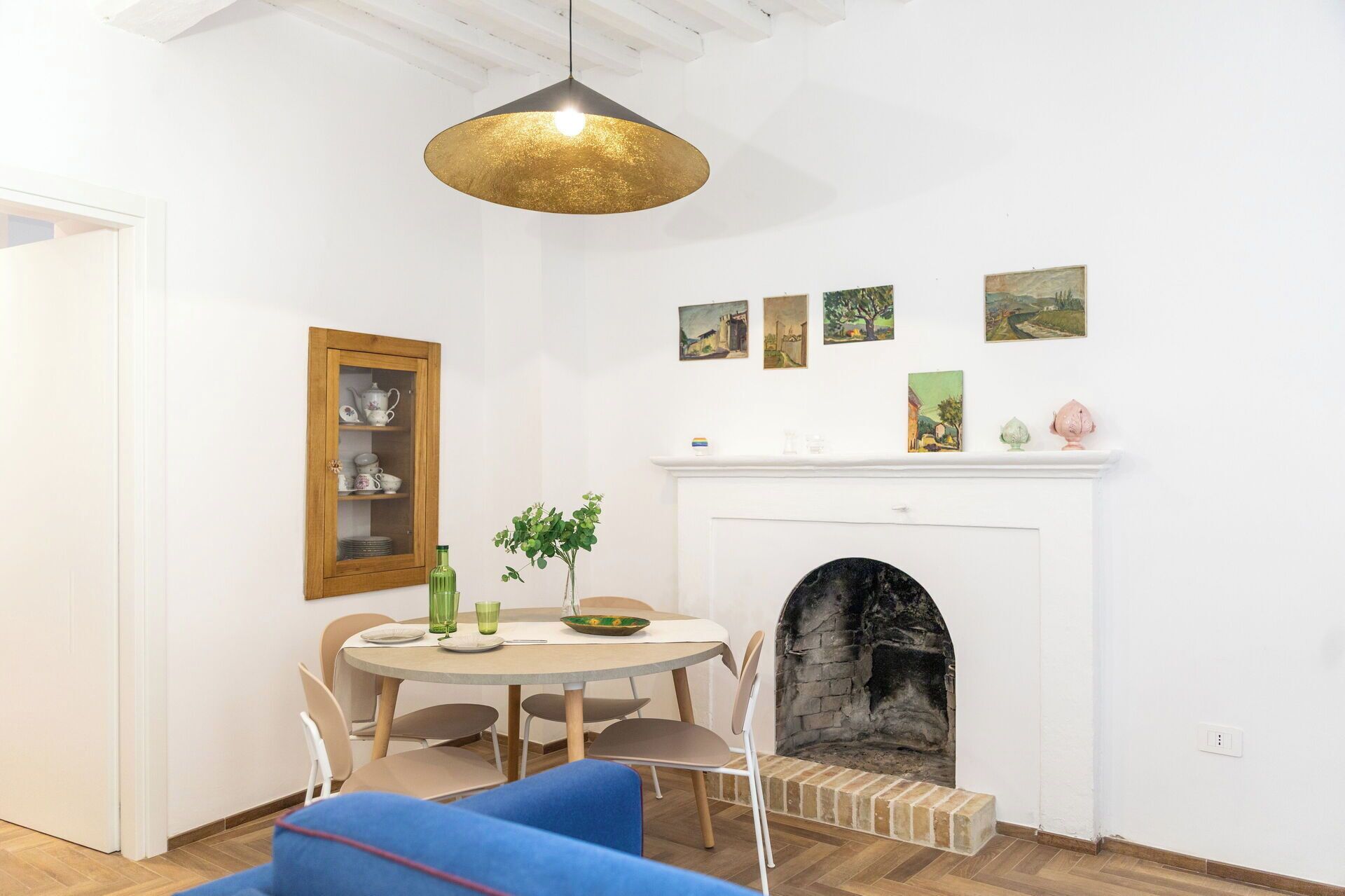 A dining area with a round table, chairs, a vase with greenery, a fireplace, and a hanging pendant light.